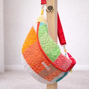 May include: A colorful, quilted crossbody bag with a curved shape. The bag features a patchwork design with orange, green, red, and white fabrics. It has a red strap and a yellow zipper. The bag is hanging on a wooden post.