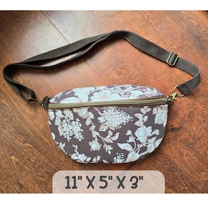 May include: A crescent-shaped fanny pack featuring a dark brown fabric with a light beige floral pattern. The bag has a brown zipper closure, an adjustable dark brown strap with metal hardware, and dimensions of 11 inches by 5 inches by 3 inches.