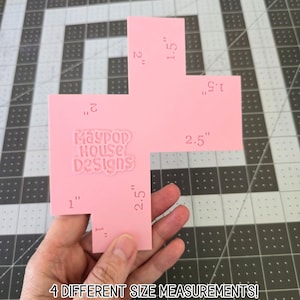May include: A pink, cross-shaped craft template with four different size measurements in inches. The template has the text "Maypop House Designs" and the text "4 DIFFERENT SIZE MEASUREMENTS!" printed on it. The template is held in a hand.