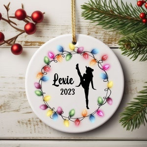 May include: White ceramic Christmas ornament with a silhouette of a dancer in a circle of colorful lights. The ornament says "Lexie 2023".