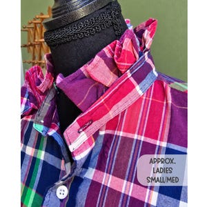 May include: A plaid shirt featuring a ruffled collar, long sleeves, and a button-down front. The fabric displays a multi-colored pattern of purple, pink, blue, and green. A white label on the lower right reads: APPROX. LADIES SMALL/MED.