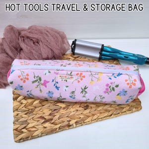 May include: A pink floral travel and storage bag with a white zipper, containing a curling iron. The bag is decorated with colorful flowers and the text "MAYWOOD HOUSE". The bag is on a woven mat.