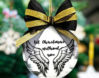 May include: A white Christmas ornament with a black bow and gold ribbon. The ornament has a pair of angel wings and the text "1st Christmas without you".