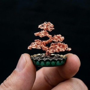 May include: A miniature bonsai tree sculpture crafted from coiled copper wire, featuring a twisted trunk and layered branches. The tree is anchored within a small, green, fluted metal bottle cap filled with dark, granular material at the base.