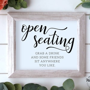 Op de afbeelding: Een wit ingelijst bord met zwarte tekst die "open seating" en "Grab a drink and some friends sit anywhere you like." luidt. Het bord staat op een witte houten achtergrond.
