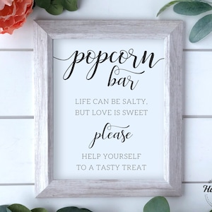 Op de afbeelding: Een wit ingelijst bord met zwarte tekst die "popcorn bar", "Het leven kan zout zijn, maar liefde is zoet", "alsjeblieft" en "Neem gerust een lekker hapje" zegt.