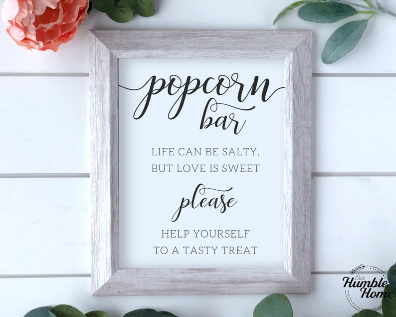 Op de afbeelding: Een wit ingelijst bord met zwarte tekst die "popcorn bar", "Het leven kan zout zijn, maar liefde is zoet", "alsjeblieft" en "Neem gerust een lekker hapje" zegt.