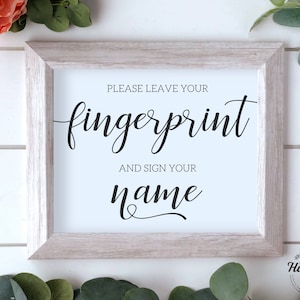 Peut inclure: Une pancarte blanche avec un cadre en bois clair, avec le texte "Veuillez laisser votre empreinte digitale et signer votre nom" en écriture cursive noire.