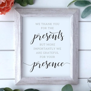 May include: A white framed sign with black text that reads "We thank you for the presents but more importantly we are grateful for your presence". The sign is on a white wood background.