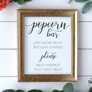 Op de afbeelding: Een gouden lijst met de tekst "popcorn bar" in zwarte cursieve letters. Het bord bevat ook de tekst "Life can be salty, but love is sweet. Please help yourself to a tasty treat." in zwarte letters.