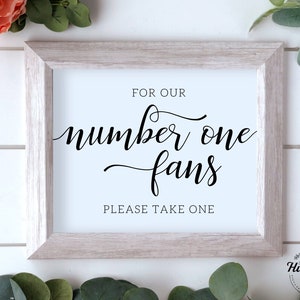 May include: A framed sign with the text "FOR OUR number one fans PLEASE TAKE ONE" in black script. The frame is light gray, and the background is white. The sign is decorated with greenery and a flower.