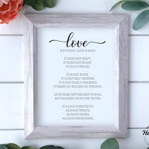 Op de afbeelding: Een ingelijste print met een witte achtergrond en zwarte tekst. De tekst luidt "liefde is geduldig, liefde is vriendelijk. Ze is niet jaloers. Ze is niet opschepperig. Ze is niet trots. Ze is niet onbeleefd. Ze is niet zelfzuchtig. Ze is niet snel boos. Ze houdt geen rekening met onrecht. Liefde verheugt zich niet over het kwaad, maar verheugt zich over de waarheid. Ze beschermt altijd. Ze vertrouwt altijd. Ze hoopt altijd. Ze volhardt altijd." De lijst is van licht hout.