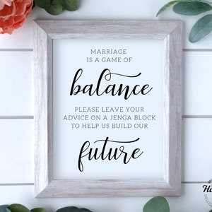 Puede incluir: Un cartel enmarcado en blanco con texto negro que dice "El matrimonio es un juego de equilibrio. Por favor, deja tu consejo en un bloque de Jenga para ayudarnos a construir nuestro futuro."