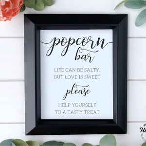 Op de afbeelding: Een zwart ingelijst bord met een witte achtergrond en zwarte tekst die "popcorn bar" luidt met de tagline "Het leven kan zout zijn, maar liefde is zoet" en "neem gerust een lekker hapje".