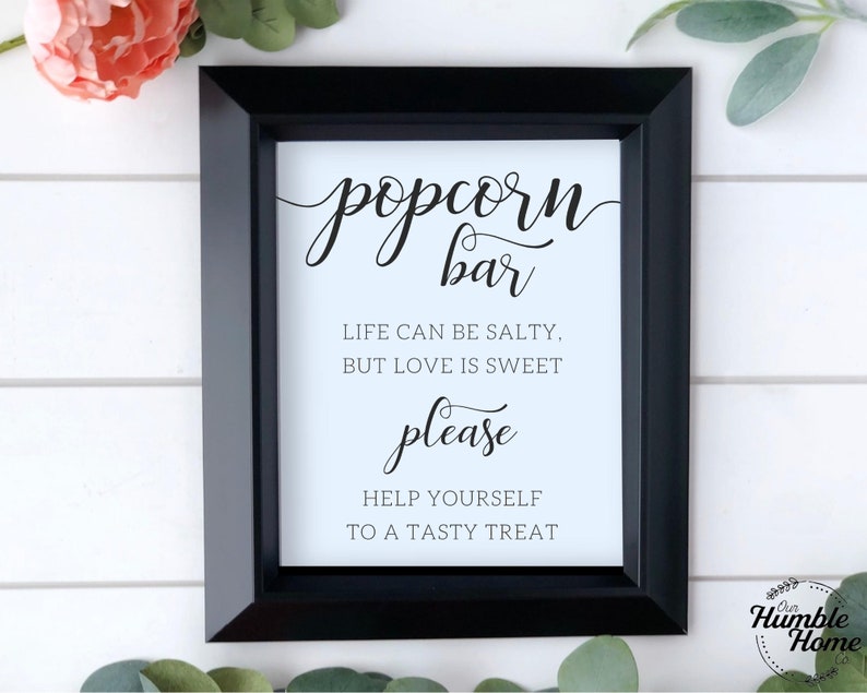 Op de afbeelding: Een zwart ingelijst bord met een witte achtergrond en zwarte tekst die "popcorn bar" luidt met de tagline "Het leven kan zout zijn, maar liefde is zoet" en "neem gerust een lekker hapje".