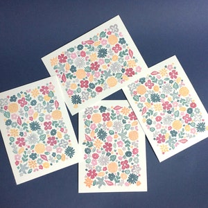 May include: Four floral notecards with a repeating pattern of various flowers in shades of pink, orange, teal, and gray. The cards are arranged in an overlapping pattern, showcasing the design from different angles. Each card features a unique arrangement of the floral motif, creating a visually appealing set of stationery.