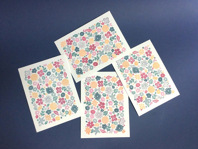 May include: Four floral notecards with a repeating pattern of various flowers in shades of pink, orange, teal, and gray. The cards are arranged in an overlapping pattern, showcasing the design from different angles. Each card features a unique arrangement of the floral motif, creating a visually appealing set of stationery.