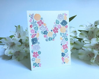May include: A white card with the letter "M" formed by a pattern of colorful flowers and leaves. The card is surrounded by white flowers.