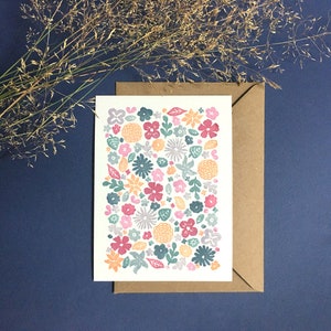 May include: A floral greeting card features a densely packed pattern of various flowers in muted pink, teal, and orange hues. The flowers are rendered in a simple, block-printed style against a white background. The card is displayed within a kraft brown envelope. The overall aesthetic is charming and understated, suitable for various occasions.