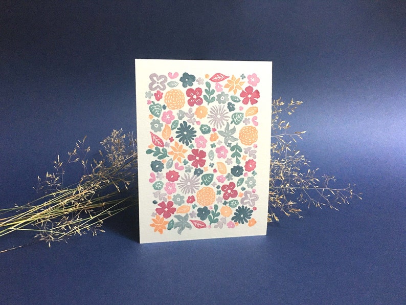 May include: A greeting card features a densely packed pattern of various flowers in shades of pink, orange, teal, and purple. The flowers are stylized and have a slightly textured appearance, suggesting a print technique like letterpress or block printing. The card's background is a light cream or off-white color, providing a contrast to the vibrant floral design. The overall style is simple, modern, and slightly whimsical, suitable for a variety of occasions.