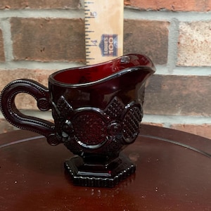 Può includere: Una lattiera in vetro rosso scuro con un motivo pressato ornato da cerchi e rombi, un manico curvo e una base esagonale. Un righello di legno posizionato dietro la lattiera indica un'altezza di circa 9 cm.