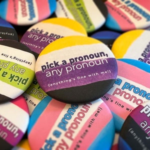 May include: A close-up of several colorful button pins with the text "pick a pronoun, any pronoun (anything's fine with me!)" on them. The buttons are arranged in a pile, with some overlapping.