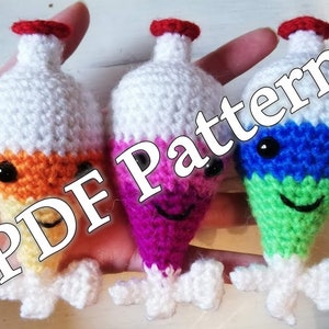 Può includere: Tre giocattoli amigurumi all'uncinetto a forma di becher con liquidi di diversi colori. I becher sono bianchi con i tappi rossi e hanno occhi neri e bocche sorridenti. I liquidi sono arancioni, rosa e verdi. Il testo "PDF Pattern" è sovrapposto all'immagine.