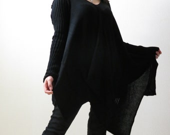 May include: A black long-sleeved sweater with a draped asymmetrical hemline. The sweater has a V-neck and ribbed sleeves. The model is wearing black pants and white sneakers.