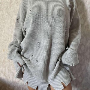 May include: A gray distressed oversized sweater with a crew neck and long sleeves. The sweater has several holes throughout the fabric.