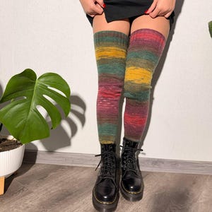 May include: Knee-high socks in a colorful striped pattern of red, yellow, green, and purple. The socks are worn with black combat boots. The boots have a shiny finish and lace-up closure. The background is a plain white wall and wooden floor.