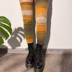May include: Over-the-knee socks in shades of olive green, orange, and beige. The socks are worn with black lace-up boots. The socks have horizontal stripes of color. The boots have a thick sole.