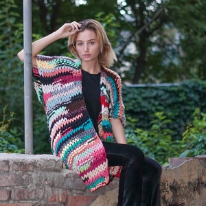 May include: A woman wearing a colourful striped crochet cardigan over a black top and black trousers. She is sitting on a brick wall with a green leafy background.