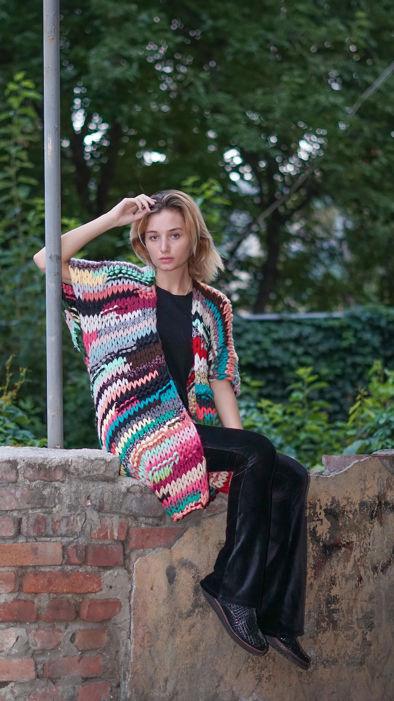 May include: A woman wearing a colourful striped crochet cardigan over a black top and black trousers. She is sitting on a brick wall with a green leafy background.