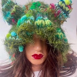 May include: A textured, fuzzy green knit hat featuring two prominent ear-like protrusions at the top. The hat is crafted from a mix of olive green, lime green, and bright turquoise yarn, accented with small pops of pink and yellow throughout the knit.