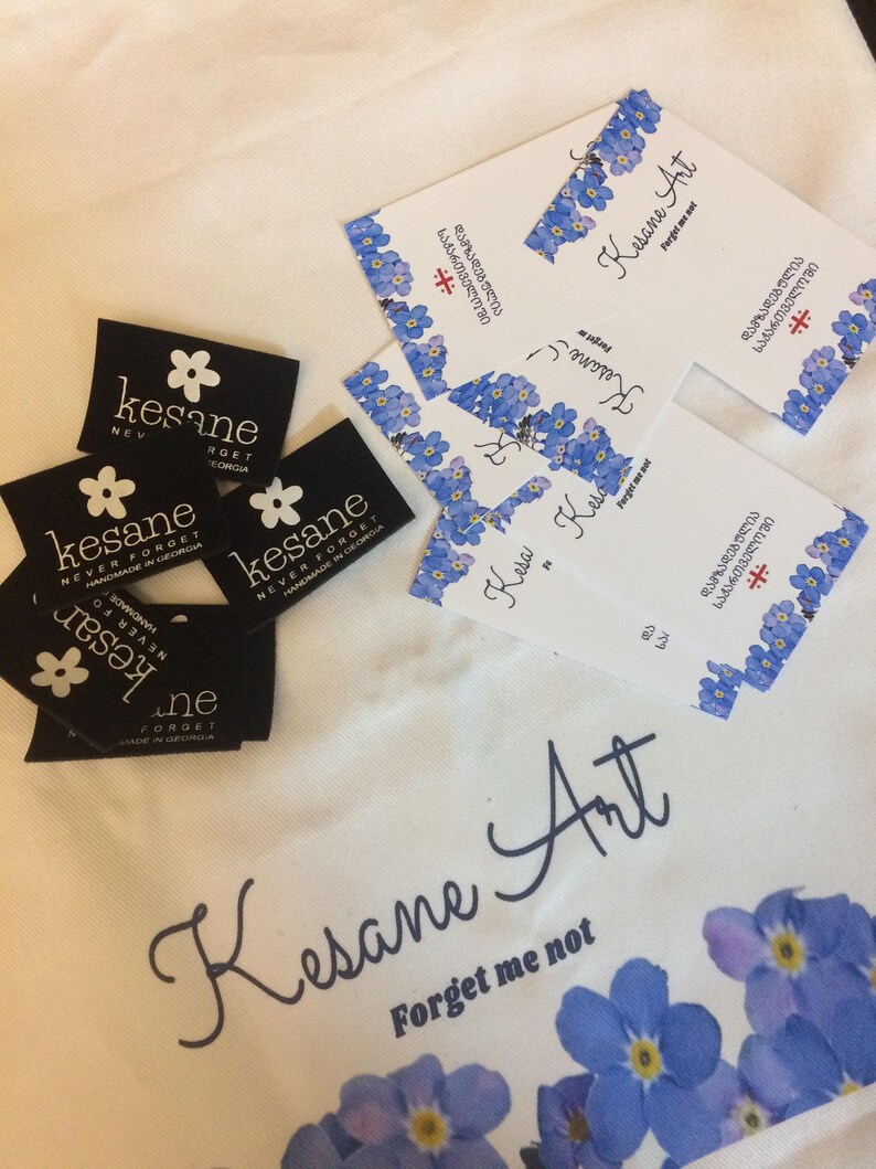May include: A white fabric bag with a blue floral pattern and the text "Kesane Art Forget me not". The bag has a black label with the text "Kesane Never Forget Handmade in Georgia".