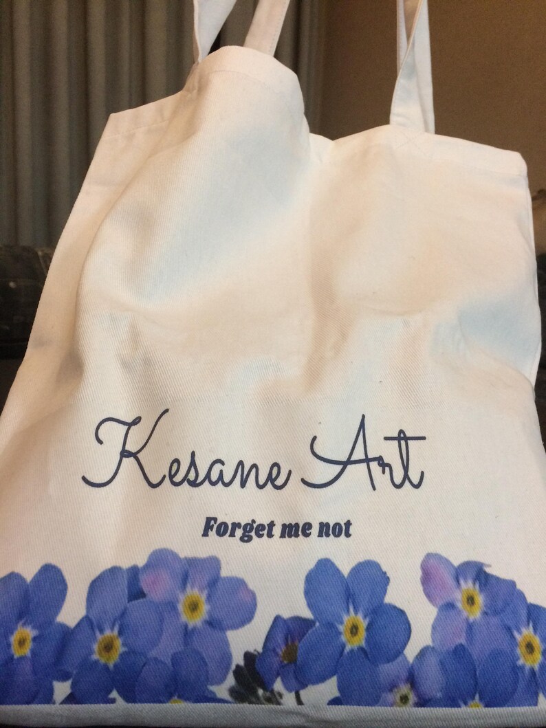 May include: White canvas tote bag with blue forget-me-not flowers printed on the bottom. The bag has blue text that says "Kesane Art" and "Forget me not".