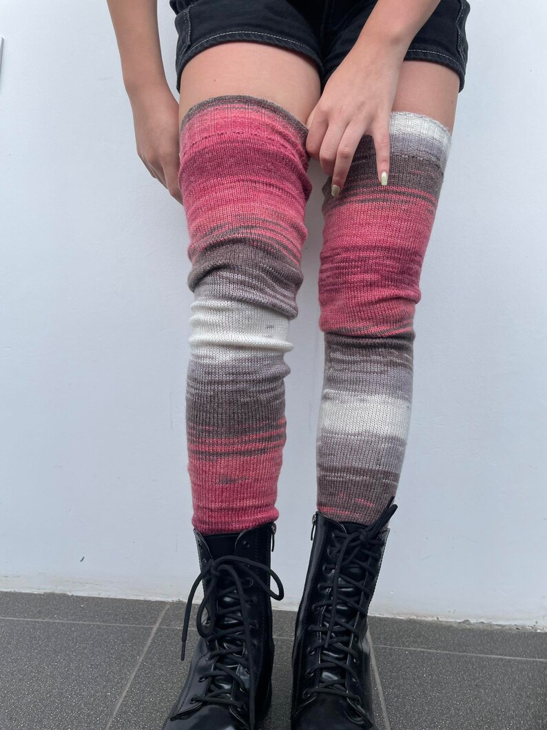 May include: A pair of long, striped leg warmers in shades of pink, brown, and white. The leg warmers are made of a soft, knitted fabric and are worn over black shorts and black boots.