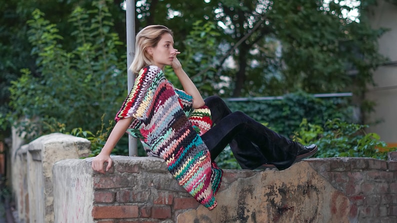 May include: A woman wearing a colourful crocheted poncho and black trousers sits on a brick wall. The poncho features a range of colours, including red, green, blue, and yellow.