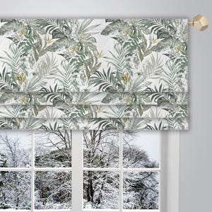 May include: A white roman shade with a green and yellow tropical leaf pattern. The shade is hung on a window with a view of a snowy landscape.