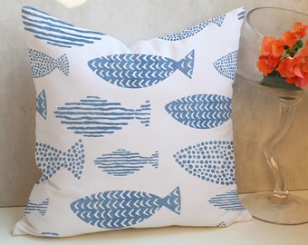 Puede incluir: Cojín cuadrado blanco con un patrón de peces azules. Los diseños de peces incluyen puntos, rayas y patrones de chevrón. El cojín está sobre una superficie blanca, con un jarrón de vidrio con flores naranjas en el fondo.