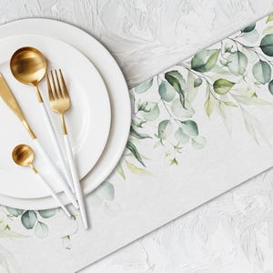 Puede incluir: Vista aérea de una mesa con platos blancos y cubiertos dorados. Los cubiertos incluyen una cuchara, un cuchillo, un tenedor y una cuchara pequeña. Un camino de mesa blanco con hojas y ramas de eucalipto verdes está a la derecha.