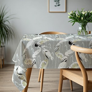 Puede incluir: Un mantel gris con un patrón repetido de garzas blancas y verde oliva y tallos de bambú. El mantel está sobre una mesa de madera. Un jarrón con flores blancas está en el fondo.