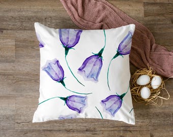 May include: White square pillow with a watercolor floral pattern of purple bellflowers and green stems. The pillow is on a wooden surface, with a pink scarf and a nest with eggs in the background.