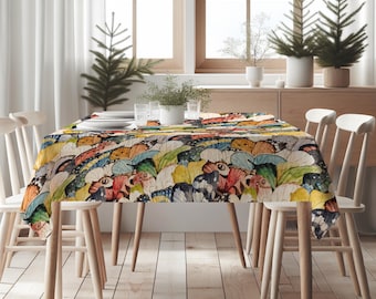 May include: A rectangular tablecloth with a vibrant butterfly wing pattern in shades of yellow, orange, blue, and red. The tablecloth is set on a wooden table with white chairs. The room has natural light and decorative plants.