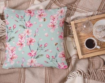 Puede incluir: Cojín cuadrado con un estampado floral de flores rosas y hojas verdes sobre un fondo verde azulado claro. El cojín forma parte de un tema de decoración del hogar, con una bandeja de madera, café y pasteles en el fondo.