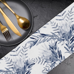 Könnte beinhalten: Ein schwarzer Teller mit goldenem Besteck, darunter eine Gabel, ein Messer und ein Löffel, liegt auf einer dunklen Oberfläche. Ein weißer Tischläufer mit blauem Blumendesign mit Blättern und Blumen ist ebenfalls zu sehen.