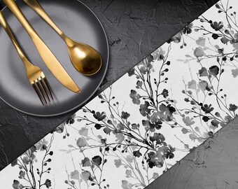 May include: A black plate with gold cutlery, including a fork, knife, and spoon, sits on a textured black surface. A floral patterned runner in black and white adds a decorative touch to the table setting.