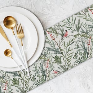 Puede incluir: Un plato blanco con cubiertos dorados y un camino de mesa decorativo. El camino de mesa presenta un diseño botánico con hojas verdes, bayas rojas y pequeños pájaros. Los cubiertos tienen mangos blancos.