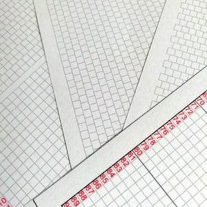 May include: Multiple sheets of white paper featuring a printed grid pattern of small gray rectangles. One sheet displays a vertical column of red numbers ranging from 10 to 15 and 70 to 90, marking the edge of the grid layout on the white paper surface.