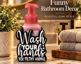 May include: A black soap dispenser holder with white text that reads Wash YOUR hands YOU FILTHY ANIMAL. The holder contains a pink soap bottle with a pump top. The text is written in a mix of cursive and block lettering on the front surface.